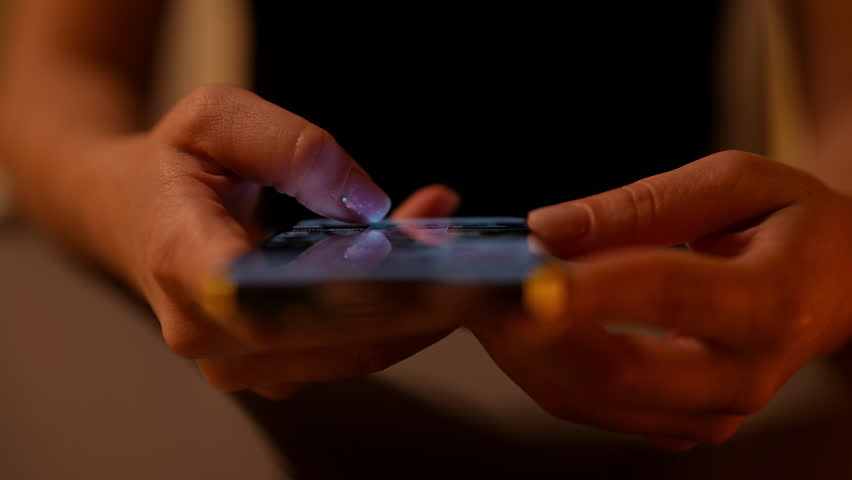 Closeup top view of woman using smartphone in dark room, browsing internet and scrolling screen with finger. Close-up of female hand scrolling using mobile phone social media in dimly lit setting.
