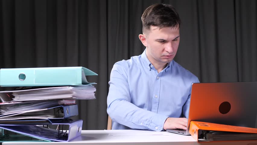 Stressed office worker looking at laptop with folders and paperwork