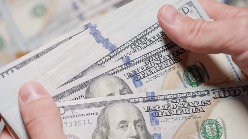 A close-up of a hand flipping through a bundle of hundred-dollar bills with scattered dollar notes blurred in the background, creating a sense of movement and wealth.