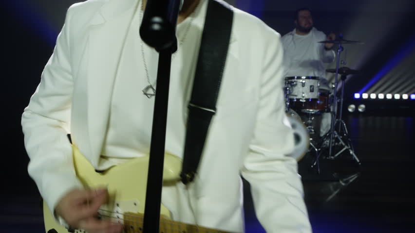 Singer playing guitar while singing with band in event. Drummer playing music on drumset in background. Musicians are performing on illuminated stage at night.