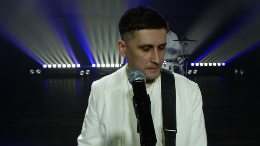 Portrait of enthusiastic pop singer singing while playing guitar. Drummer playing music on drumset in background on illuminated stage. Performers performing in musical event at night.