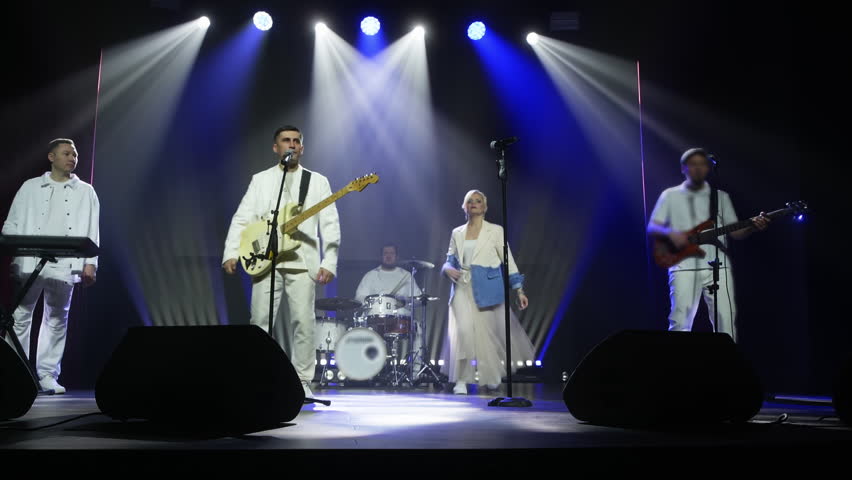 Male and female singers performing with band. Musicians playing instruments on illuminated stage at event. Artist enjoying musical show together at night.