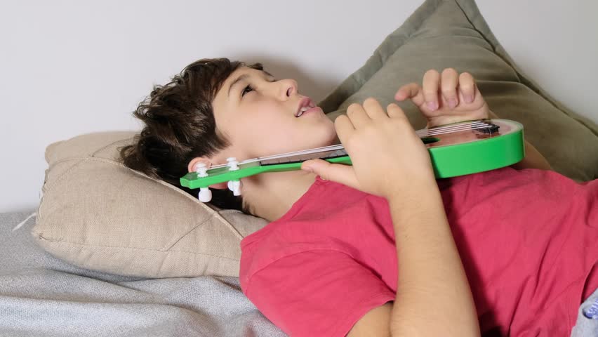 A young boy rests on pillows, holding a ukulele close as he appears deep in thought or softly singing. This image highlights the calming and inspiring role of music in personal moments.