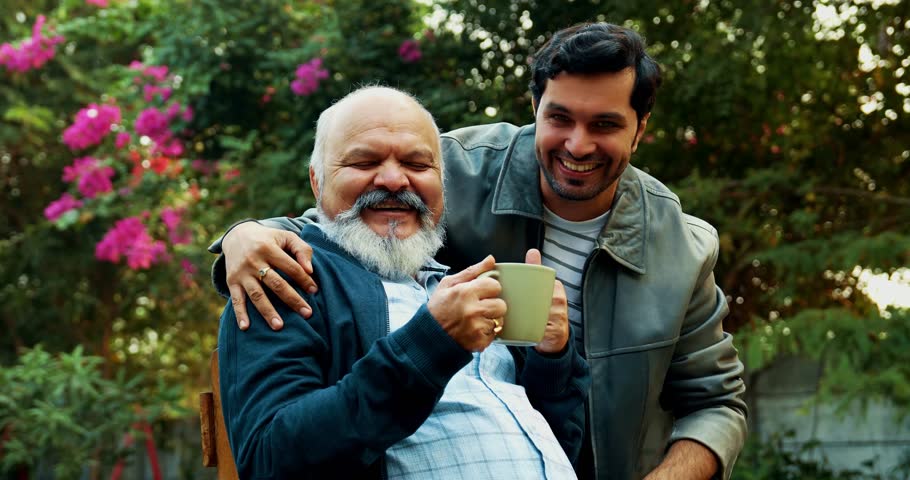 Indian Asian Happy Old Aged Father Sitting on Chair in Home Garden, Young Son Hugs Him from Behind, Both Smiling at Camera, Celebrating Father-Son Bonding and Love