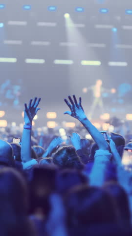 Vertical video: Back view happy people crowd on blurred rock band stage background in blue spotlights light. People move, dance, wave arms, film performance of favorite musical group concert on phones