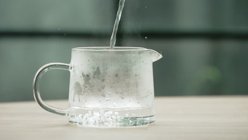Close up slow motion pouring hot boiling water into transparent glass kettle on the table of a glazed veranda