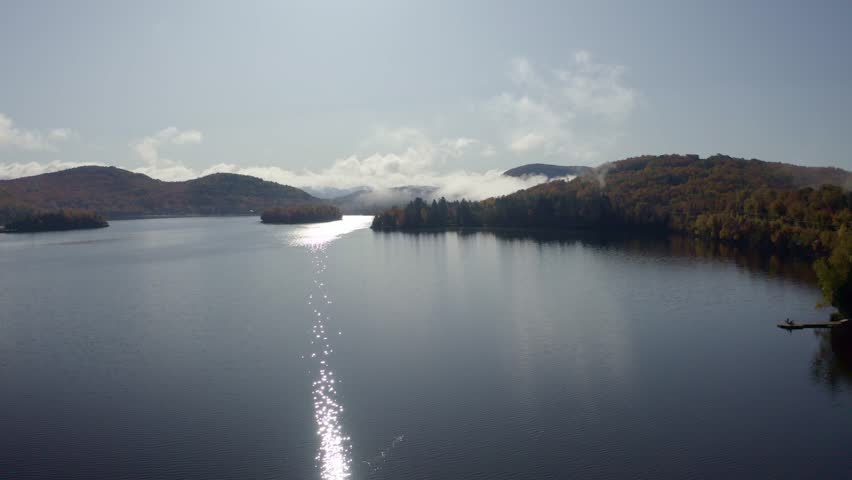 A peaceful lake reflecting the sparkling sunlight, surrounded by vibrant autumn hills partially cloaked in mist, creating a serene and captivating landscape.