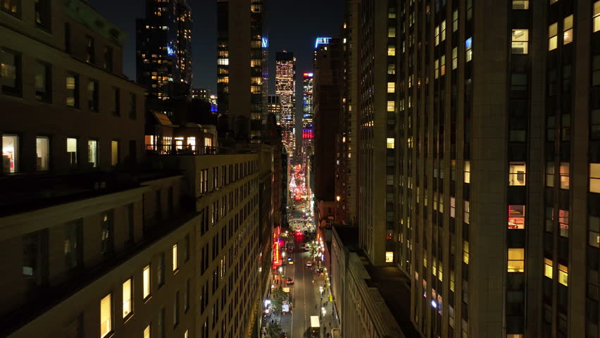 Camera glides over Midtown Manhattan, New York City street at night, capturing cars driving and residential building windows glowing with warm lights of apartments interiors