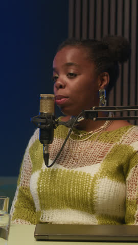 Vertical medium shot of young confident Black woman speaking in microphone and sharing her thoughts during podcast recording in studio with professional environment