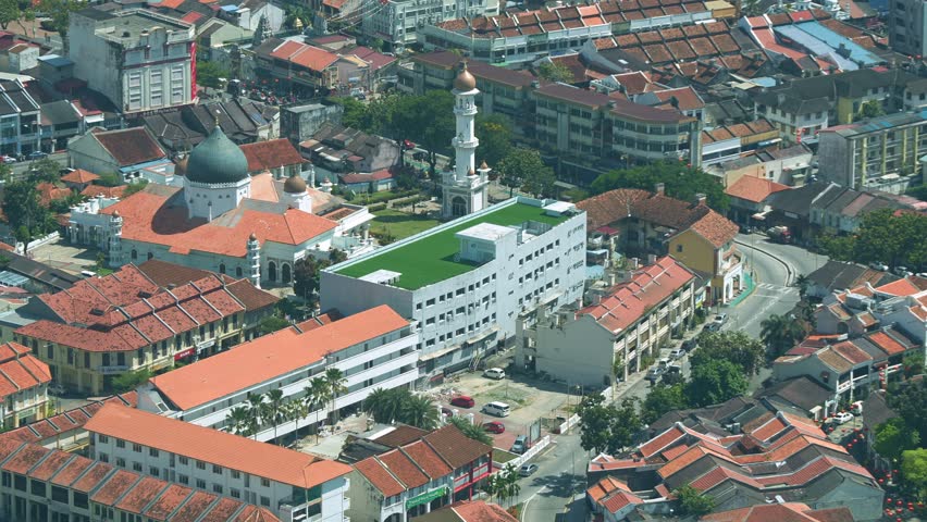 An aerial view of Penang, Malaysia, highlights Kapitan Keling Mosque with a green dome amidst a mix of colonial and modern buildings, reflecting the city