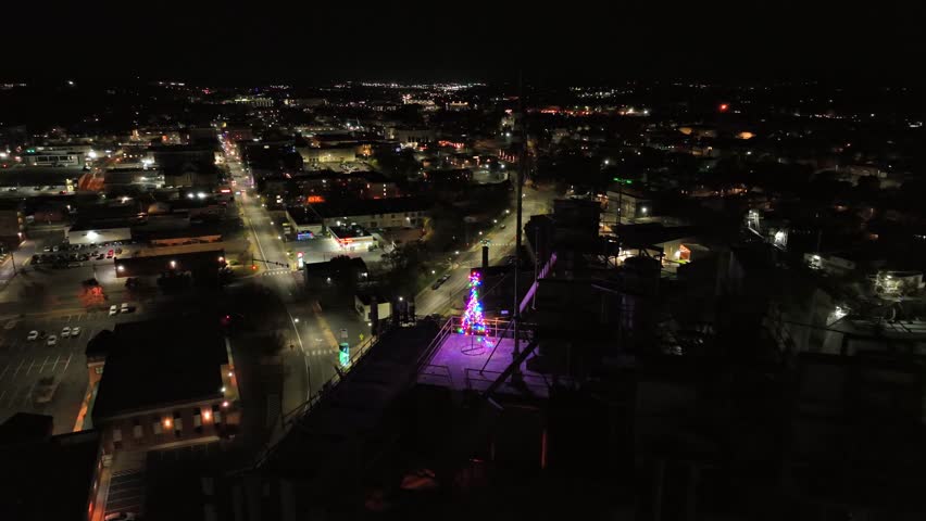 Lighting Christmas tree and american flag on top of high tower at night. American suburb area with illuminated main street. Aerial passing by flight. Wide shot.