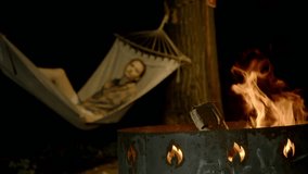 Young woman enjoys rocking and warming herself in hammock at night, lying next to brazier with bright flames from campfire escaping. Close-up - Powered by Shutterstock - Get 15% off with code: PIKWIZARD15