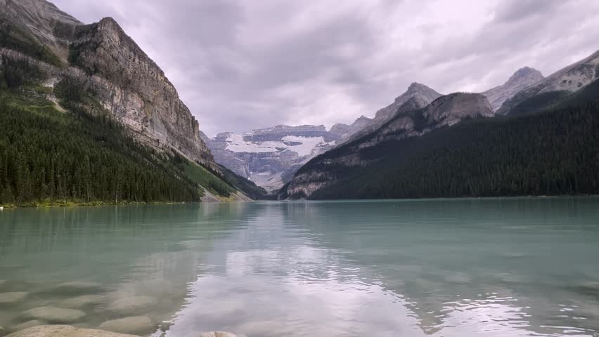 Serene view of Lake Louise turquoise waters reflecting surrounding majestic mountains and glaciers in Banff National Park, Alberta, Canada, Low angle water surface point of view