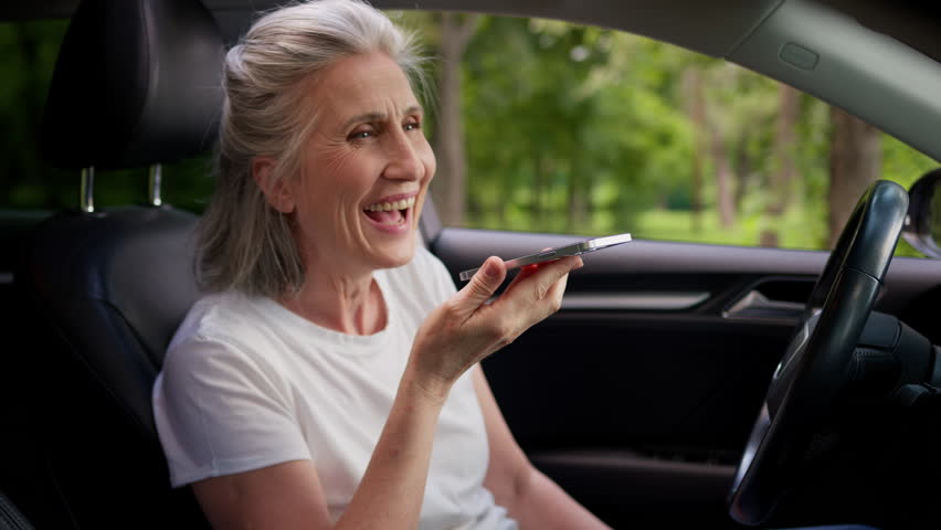 Happy smiling Caucasian elderly senior mature female woman laughing auto car driver pensioner inside automobile use mobile phone online app talking call recording voice message smartphone speakerphone