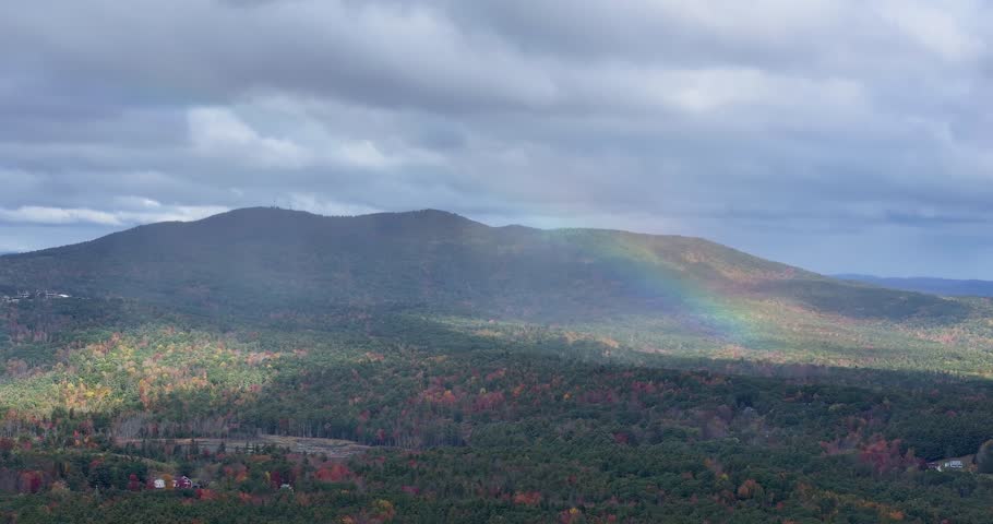 A rainbow over a forest with mountains under a cloudy sky, wide aerial view