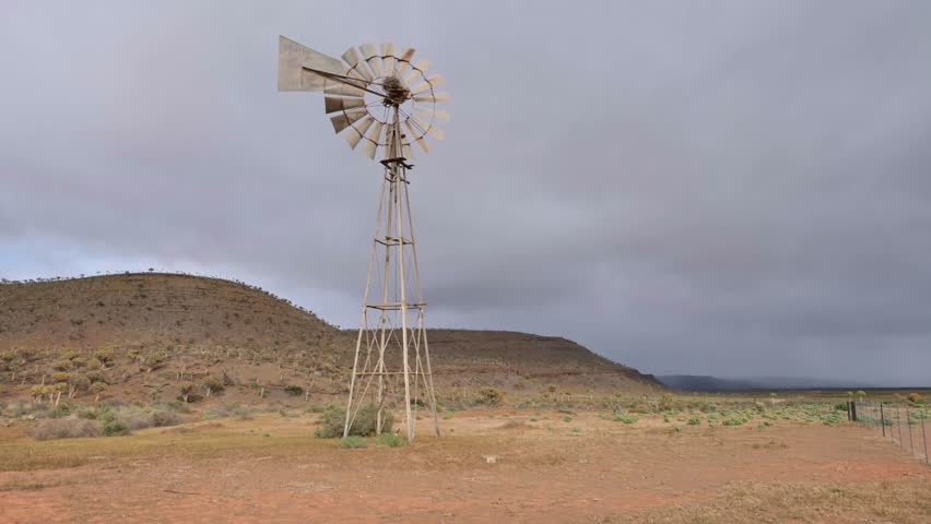 Windpump standing in arid Karoo landscape under cloudy skies with distant hills visible