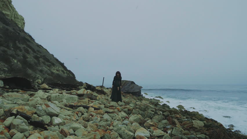 A solitary female figure in dark clothing walks along a rocky shoreline, with waves crashing nearby. A metaphor of loneliness and solitude, a concept of thoughtfulness and contemplation