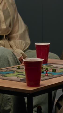 Vertical slowmo of four unrecognizable friends toasting with plastic red cups while playing tabletop game together in modern apartment