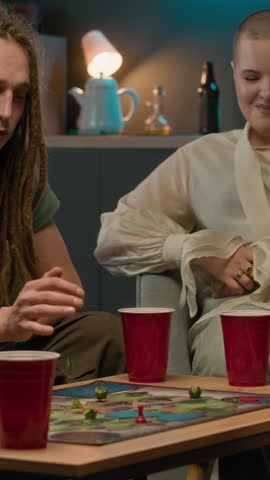 Vertical medium shot of group of young perople sharing atmosphere with drinks in red plastic cups and tabletop games, gathering together in modern apartment in evening