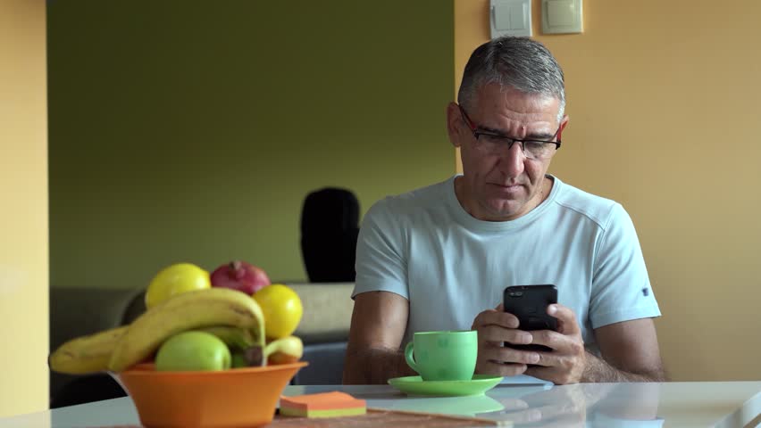 Morning Break: Chatting, Coffee, and Relaxation. A man at a table with coffee and fresh fruit texts on his phone, sips coffee, and gazes out the window. Scenes of relaxation and morning routines.