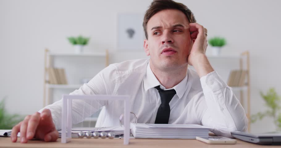 Bored exhausted company employee sitting at work desk and looking at Newton's cradle pendulum. Burnt out manager feeling unmotivated and uninspired, trying to find energy to continue his tasks.