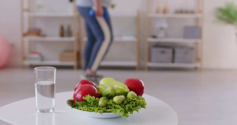 Glass of water and fresh vegetables on plate on a table with plus size, fat woman exercising at home on background. Her commitment to diet and workout training emphasizes a healthy lifestyle.
