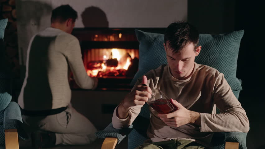 A young man sits in an armchair near a glowing fireplace, holding a bottle of strong alcohol, as another man in the background adds firewood to the fire