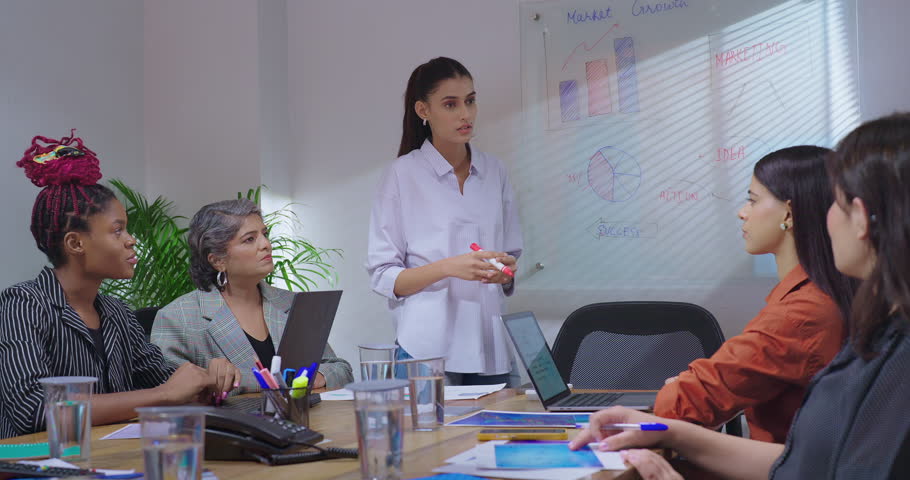 Indian young adult business woman leader show sales growth chart on white board discuss market graph data work place. Diverse female colleague listening task instruction share project ideas job office