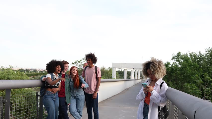 Group of four multi ethnic students leaving a classmate alone while using a smartphone on a bridge highlights themes of exclusion and social dynamics among youth
