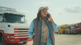 Front camera view of African American woman casually standing in parking lot for truck drivers. Talking with someone on smartphone. Remote communication concept. Lorry in background. - Powered by Shutterstock - Get 15% off with code: PIKWIZARD15