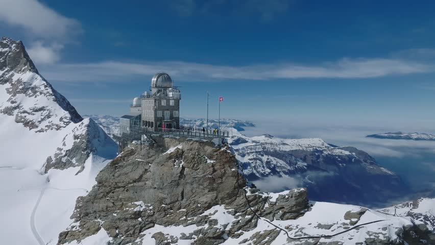 Aerial views of Sphinx Observatory with snow covered peaks and Aletsch Glacier in Grindelwald, Switzerland, under a clear blue sky with Swiss flag.