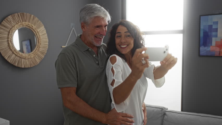 Couple taking selfie in cozy apartment shows love and happiness of middle-aged man and woman together indoors in stylish living room embracing and smiling joyfully