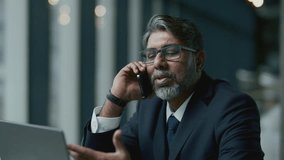 Mature Indian businessman with grey hair and beard, wearing elegant suit and glasses, sitting at cafe table with laptop on it and speaking on mobile phone - Powered by Shutterstock - Get 15% off with code: PIKWIZARD15
