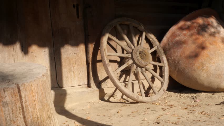 Rustic wooden wheel leaning against the wall near a large earthen pot. A weathered wooden wheel rests against a rustic wall beside an oversized earthen pot, wide shot.