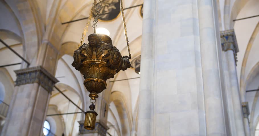 An ornate hanging lantern in a cathedral interior, featuring detailed carvings and suspended by chains, with arched ceilings and columns in the background.