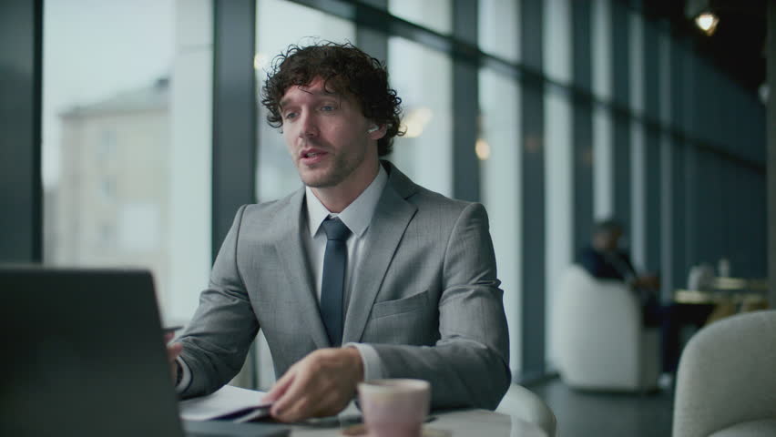 Young businessman in formal suit and wireless earphones, sitting at table in restaurant and discussing project during virtual meeting via video call on laptop