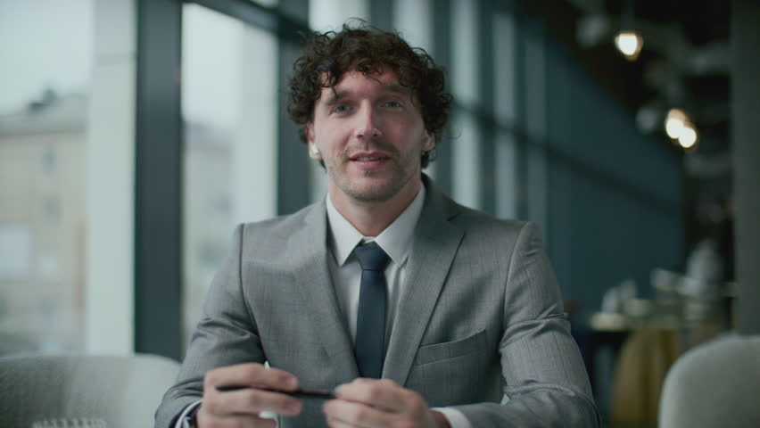 Businessman in elegant suit and wireless earbuds, sitting in restaurant, looking at camera and talking during online meeting via video call