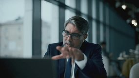 Experienced Indian businessman in wireless earphones, wearing formal suit and glasses, sitting in restaurant and talking via video call on laptop during e-meeting - Powered by Shutterstock - Get 15% off with code: PIKWIZARD15