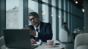 Senior Indian businessman in wireless earphones, dressed in formal attire, sitting at cafe table with coffee cup on it and having discussion via video call on laptop - Powered by Shutterstock - Get 15% off with code: PIKWIZARD15