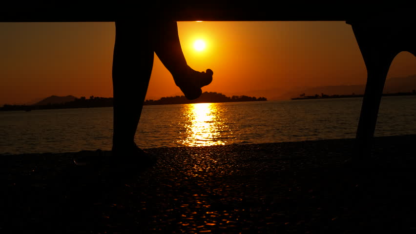 Woman on bench look at lake in dusk. A view of female legs silhouette on bench by the calm lake waves during dusk time.