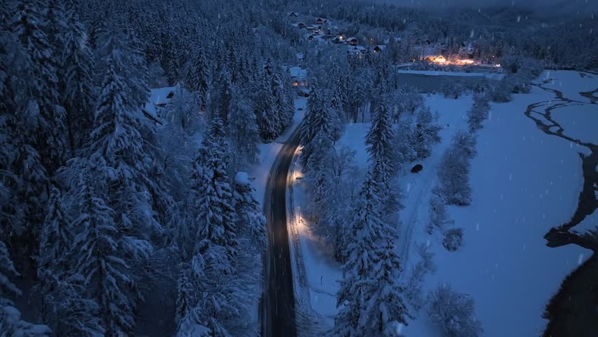 Aerial view of mountain road in fairy forest during snowfall at night. Winter fairytale. Top drone view of mountain village, houses, illumination, snowy pine trees at twilight. Beautiful Slovenia