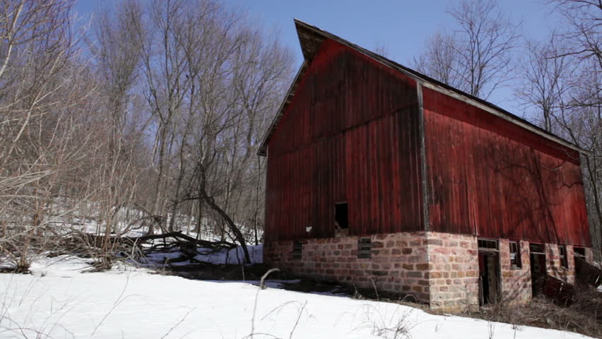 Weathered red barn of an abandoned 1910 homestead hidden in snowy woods in rural Wisconsin. The wood of the beautiful old barn is faded and textured with a stone foundation. Slow pan.