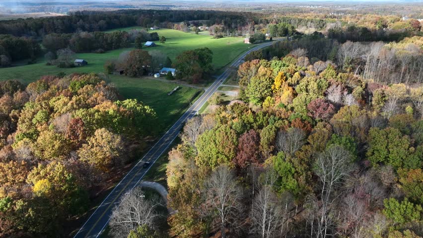 Driving cars on rural road in suburb of American town. Colorful trees and green farm fields in autumn season. Aerial flyover shot. Single family houses and barns in USA. Top down shot.