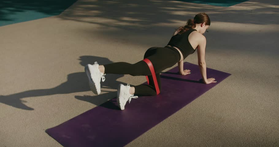 Athletic brunette female in sportswear exercising, stay in plank with resistance band and raise leg up. Sweaty woman in sportswear doing exercise on muscles of thigh, raises and lowers her leg