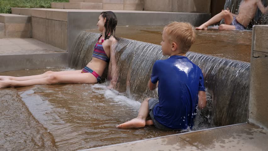A boy and a girl in wet clothes are playing with water in a fountain, they are sitting in a fountain on the ground. Children play in the fountain on a hot summer day in the city park.