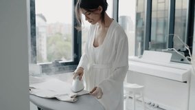 A woman in a white robe irons clothes in a bright, modern room with large windows and a piano. The scene exudes calm and elegance. Woman Ironing Clothes at Home in Bright Modern Apartment - Powered by Shutterstock - Get 15% off with code: PIKWIZARD15