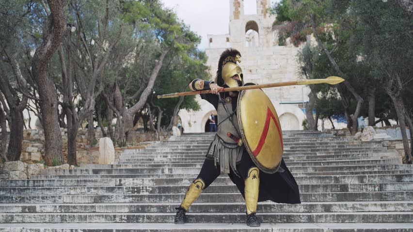 Portrait of hoplite spartan Greek soldier pose at Acropolis wearing metal helmet, protecting shield and sharp spear at Odeon of Herodes Atticus in Athens, Greece, slow motion cinematic shot