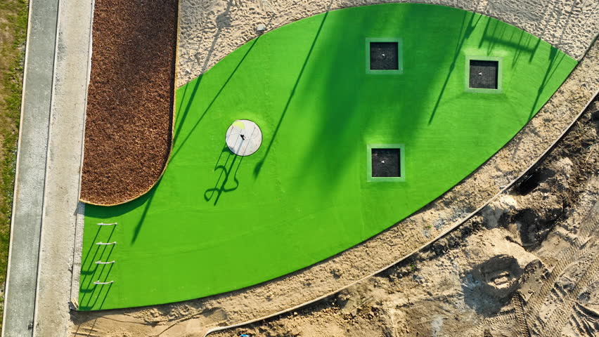 Top-down view of a green artificial playground area in the shape of a leaf with sandy surroundings, located near Gdynia.