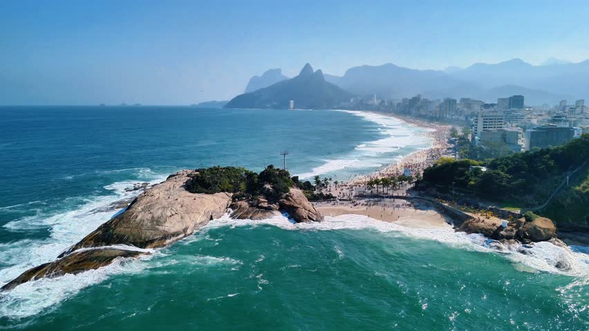 Drone flies over Pedra do Arpoador and Ipanema Beach, showcasing beautiful white sandy beach, blue ocean, palm trees, skyscrapers, and mountains in Rio de Janeiro