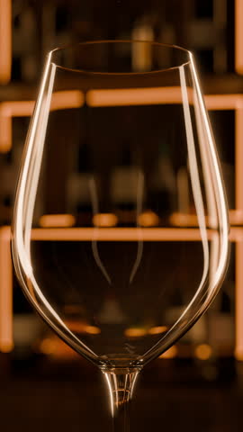 Pink or rose wine pouring in vertical slow motion inside of warm lit bar. Illuminated bar shelves with different alcohol bottles. Friday night celebration. Close up vertical shot.